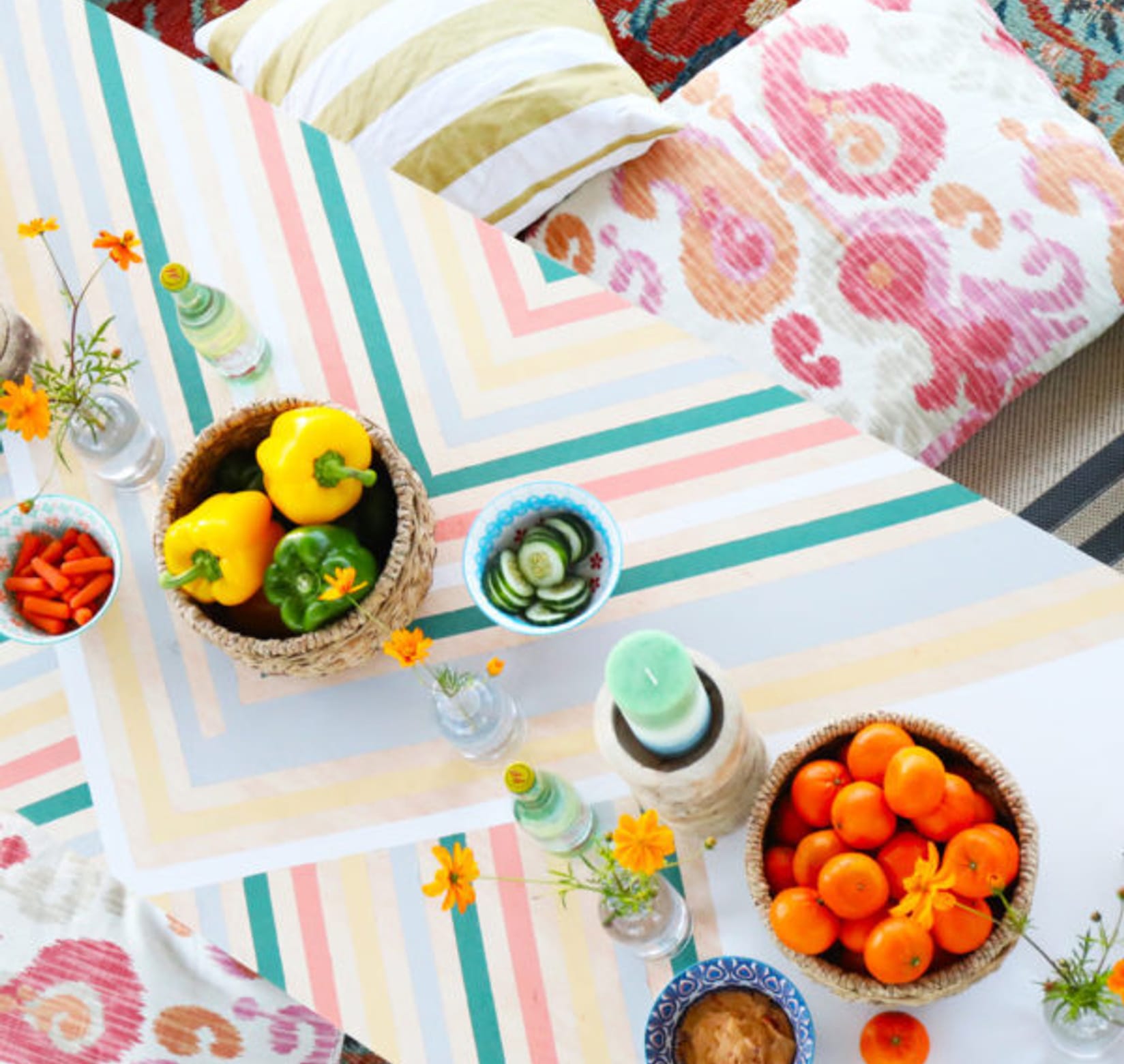 Table with V patterned lines in light pastel colors. There are peppers and clementines in bowls on the table. There are also some flowers on the table and pillows on either side.