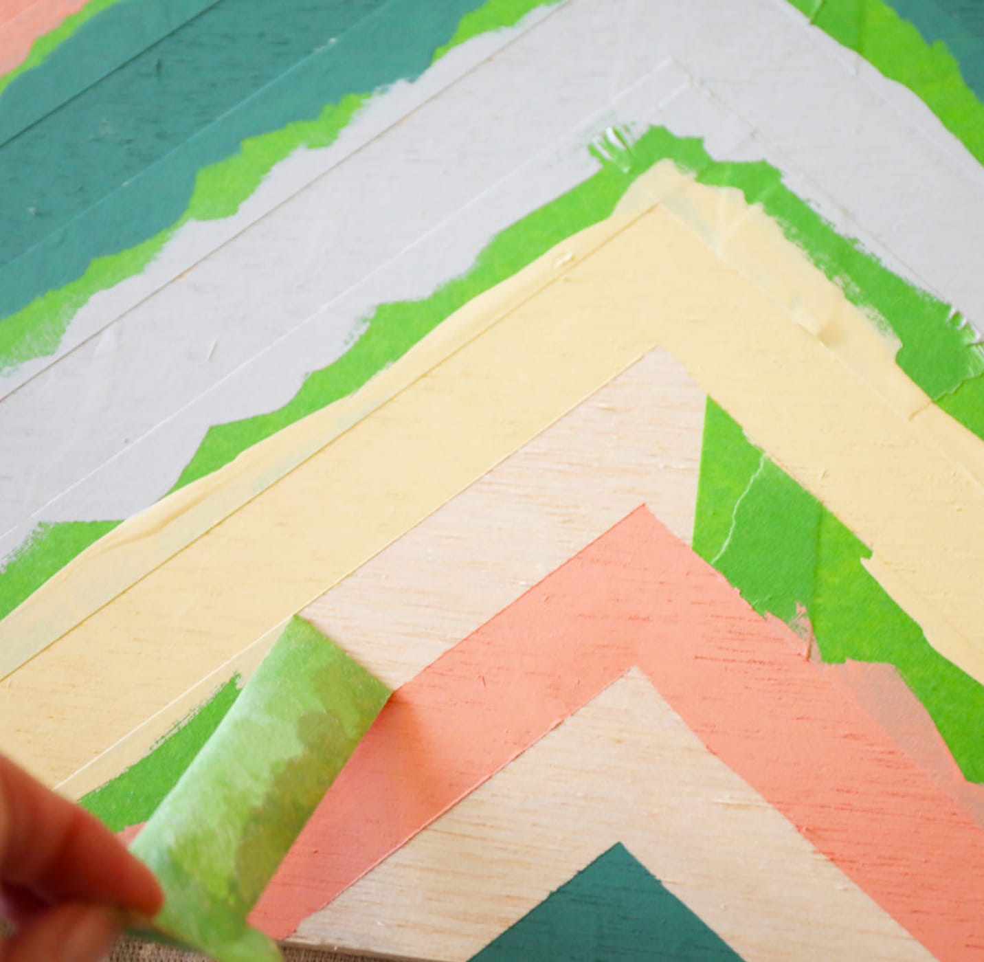 V shaped strips on Green FrogTape Multi-Surface Painter's Tape on a board with pastel colored paint. One strip of tape is being peeled off to reveal a crisp line.