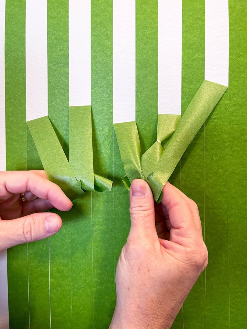 Green FrogTape Multi-Surface Painter's Tape on a wall vertically, directly next to one another. There is a set of hands pulling every other strip down off the wall