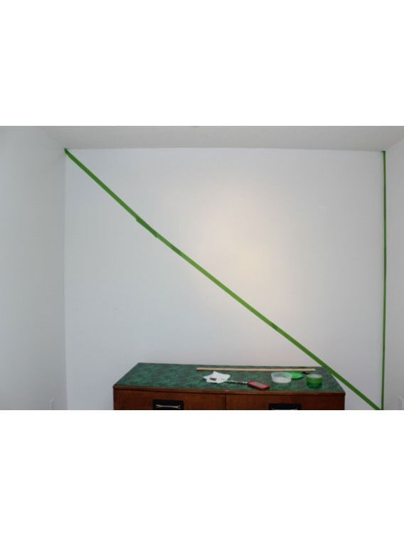 A white wall with a diagonal green tape line going across from top left to bottom right. Small wooden table with green top at the bottom of the image.