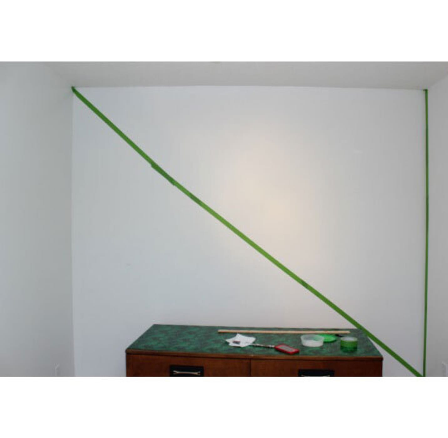 A white wall with a diagonal green tape line going across from top left to bottom right. Small wooden table with green top at the bottom of the image.