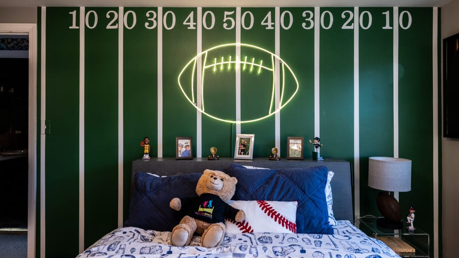 Sports bedroom with football field wall mural. There is a bed with blue and white sports themed sheets and a teddy bear on the bed. The side table on the right has a foorball lamp and there is a neon football sign hanging over the bed.