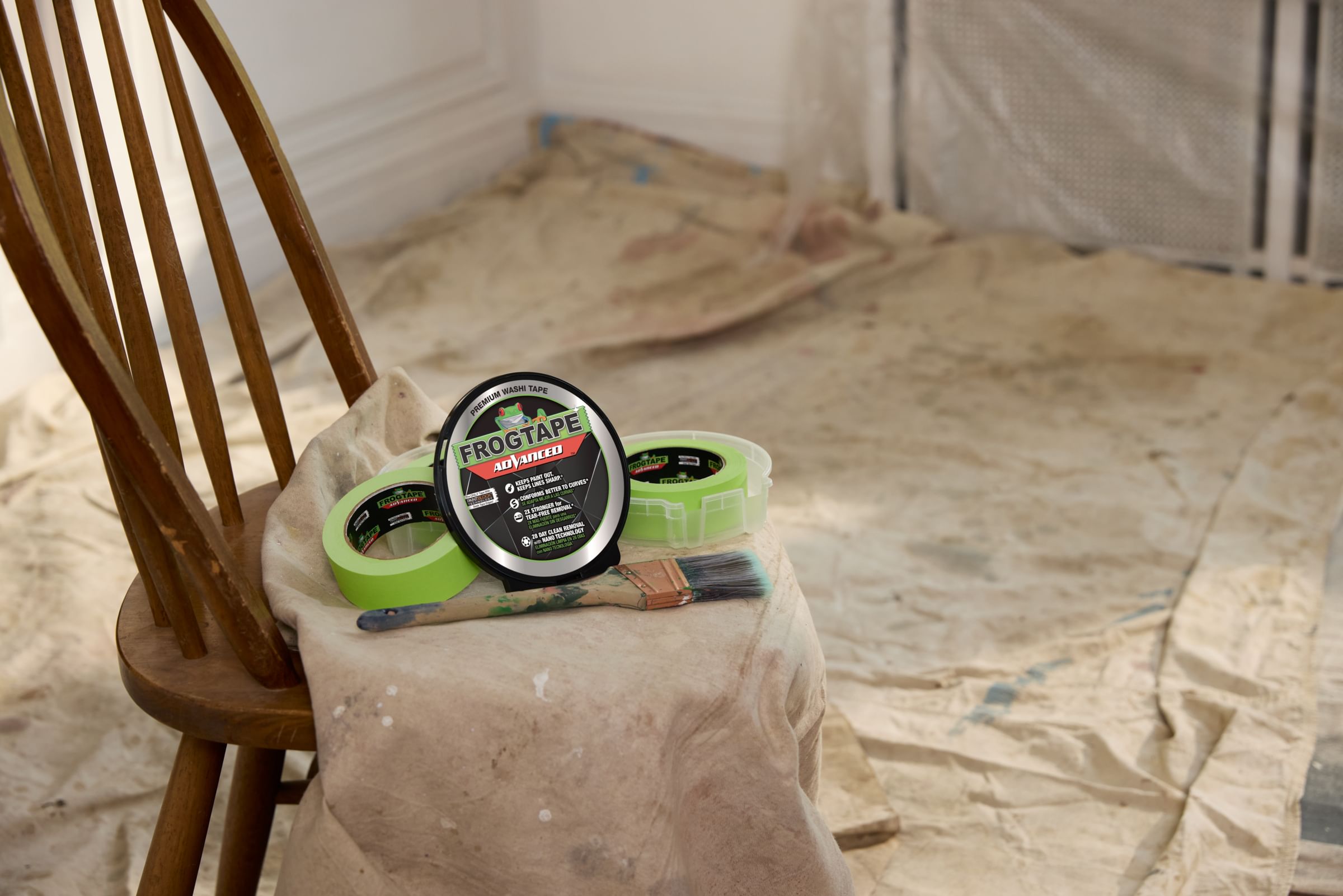 Two rolls of green FrogTape Advanced Painter's Tape in open canisters on a drop cloth on a kitchen chair with a paintbrush and the canister lid propped up. The ground in the background is covered with drop cloths