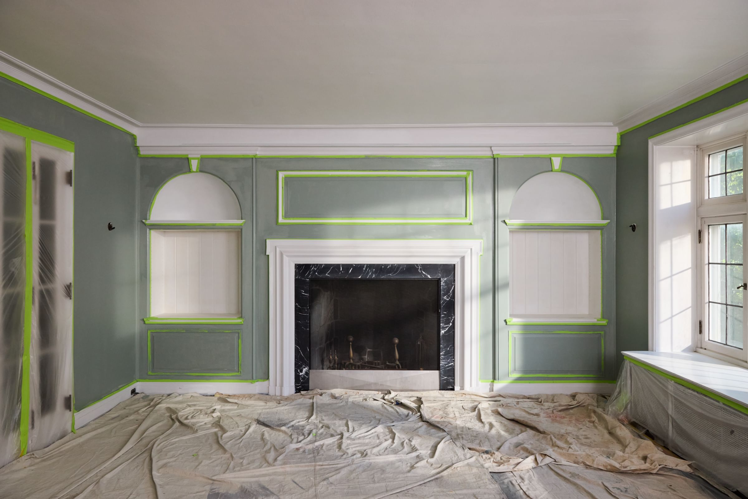 Living room with fireplace in the center of the back wall. The room is freshly painted, there are drop cloths on the floor, plastic sheeting on the doors and cabinet, and FrogTape Advanced taping off sections of the walls and ceiling.