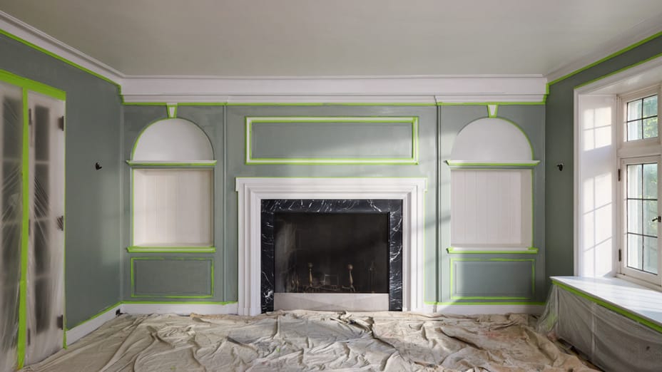 Living room with fireplace in the center of the back wall. The room is freshly painted, there are drop cloths on the floor, plastic sheeting on the doors and cabinet, and FrogTape Advanced taping off sections of the walls and ceiling.