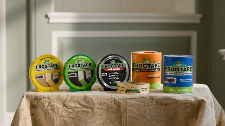 Table with Painters drop cloth and FrogTape Family of Products displayed. Right to left: Yellow FrogTape Delicate Surface Painter's Tape, Green FrogTape Multi-Surface Painter's Tape, FrogTape Advanced Painter's Tape, Beige FrogTape High Bond Exterior Painter's Tape, FrogTape Pro Grade Orange Painter's Tape, and Blue FrogTape Pro Grade Painter's Tape