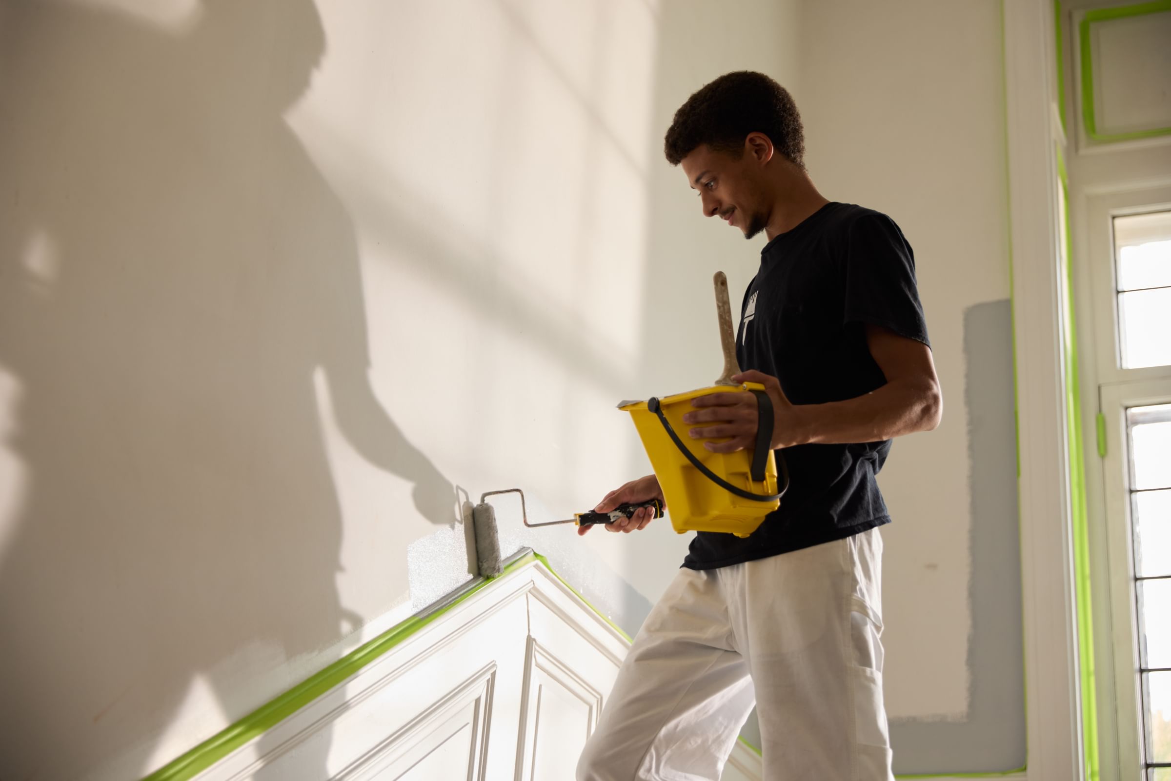 Pro Painter holding a yellow pant cup and painting the wall with a paint roller. There is Lime Green FrogTape Advanced taping off the wainscotting on the lower part of the wall.