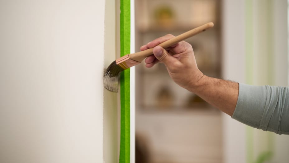 A hand with paintbrush painting a wall brown right up next to the trim of a door that is covered with Green FrogTape Multi-Surface Painter's Tape