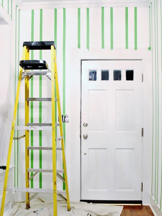 Entryway area with white door. There is a yellow ladder and white drop cloth on the ground. There are vertical strips of Green FrogTape Multi-Surface Painter's Tape taped on the walls from the ceiling to the floor.