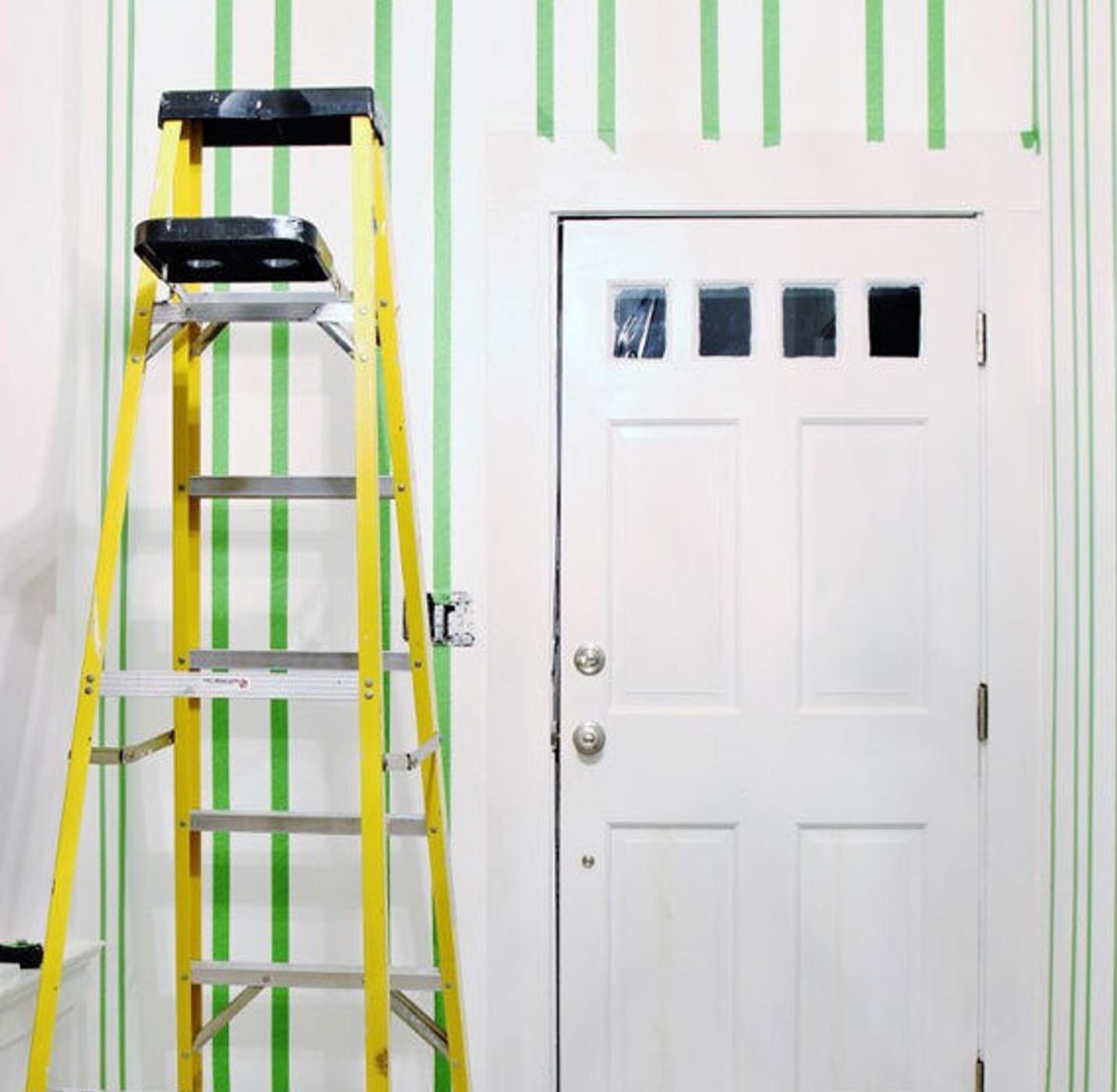 Entryway area with white door. There is a yellow ladder and white drop cloth on the ground. There are vertical strips of Green FrogTape Multi-Surface Painter's Tape taped on the walls from the ceiling to the floor.