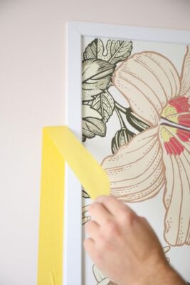 Yellow FrogTape Delicate Surface Painter's Tape being peeled off between the wall and a white frame with a painting of a flower