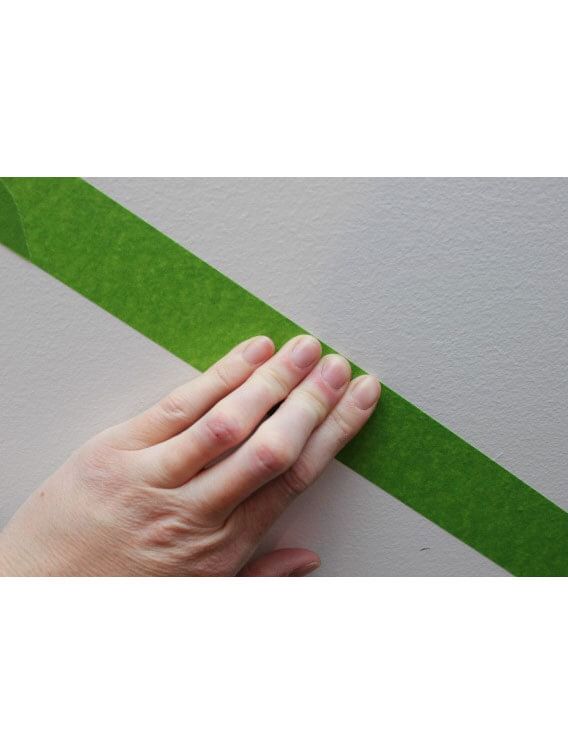 A hand firmly pressing down Green FrogTape Multi-Surface Painter's Tape on a white wall