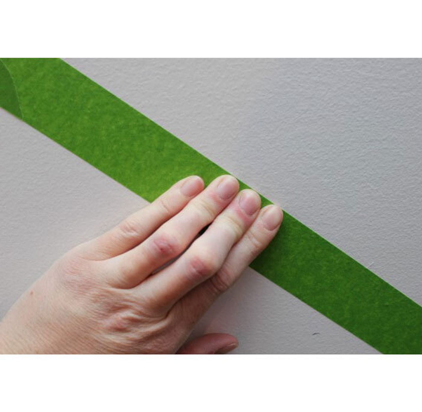 A hand firmly pressing down Green FrogTape Multi-Surface Painter's Tape on a white wall