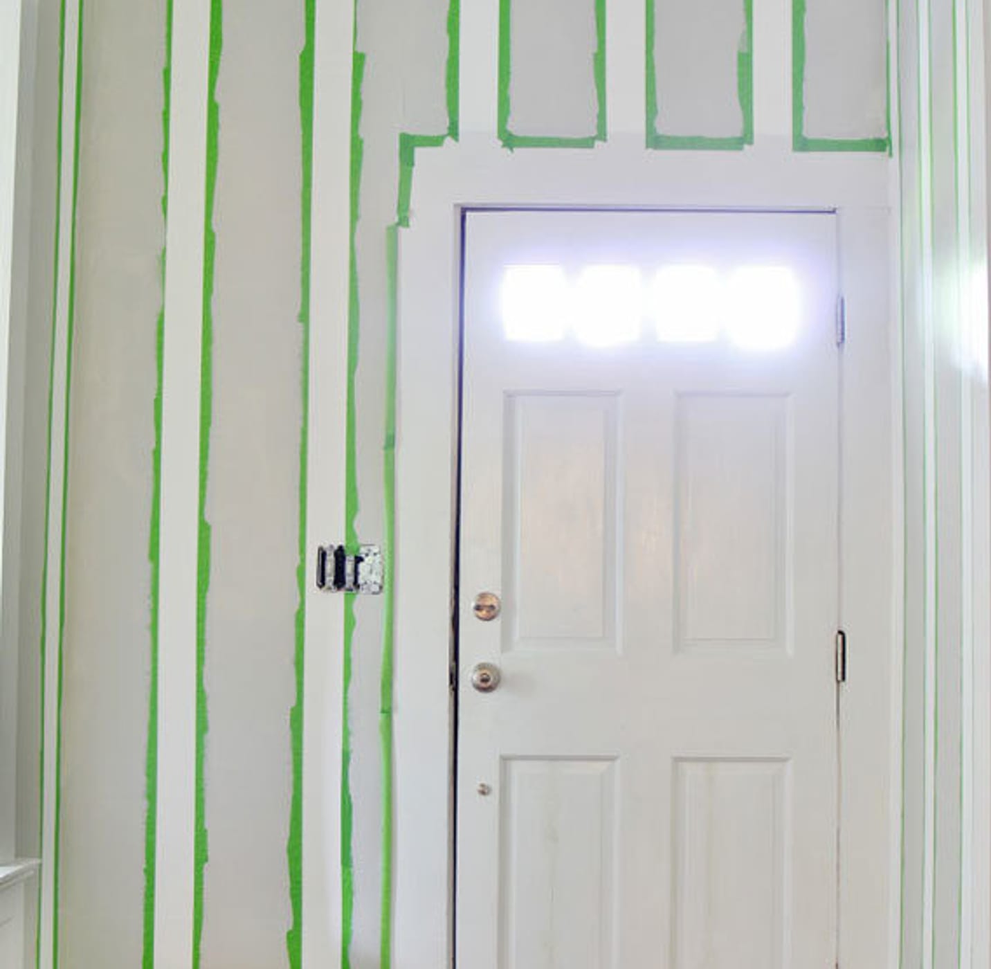 Puerta de entrada blanca. La pared está encintada del techo al suelo con cinta de pintor FrogTape Multi-Surface verde espaciada. Las franjas alternadas de la pared son de color gris y blanco.