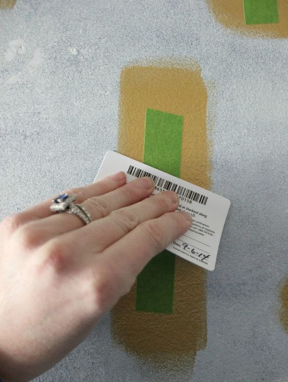A hand applying Green FrogTape Multi-Surface Painter's Tape with a credit card to a large gold rectangular paint splotch.