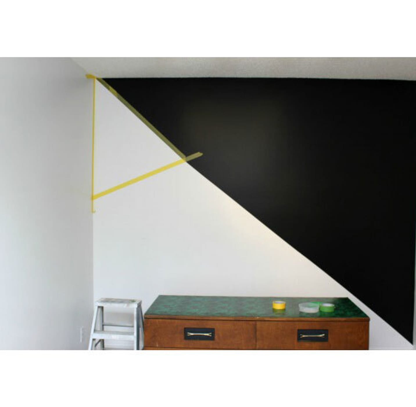 Wall cut in half by a diagonal line. Left side white with a small taped off triangle in the upper left, right side black. Small wooden table with a green top near the bottom of the image