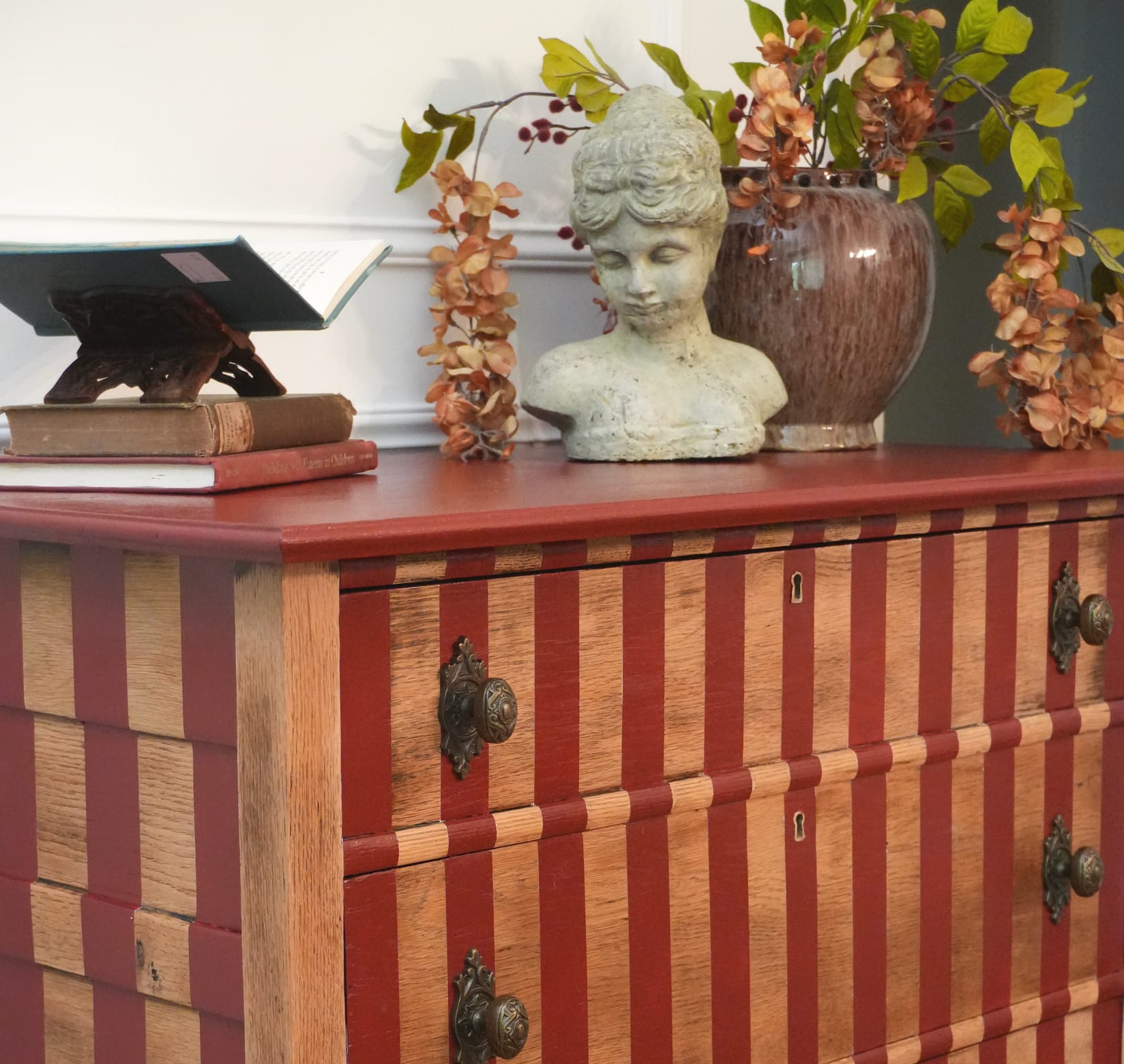 A red and oak striped dresser with a flower pot, statue bust, and some books on top