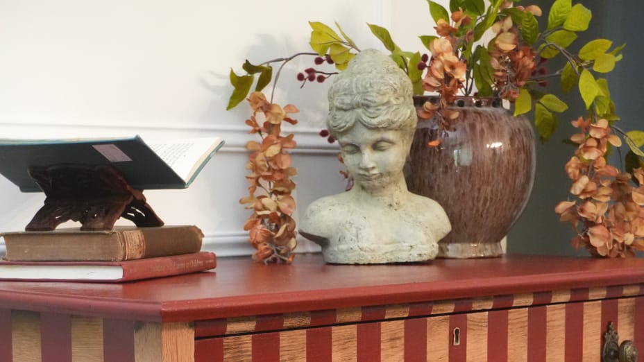 A red and oak striped dresser with a flower pot, statue bust, and some books on top