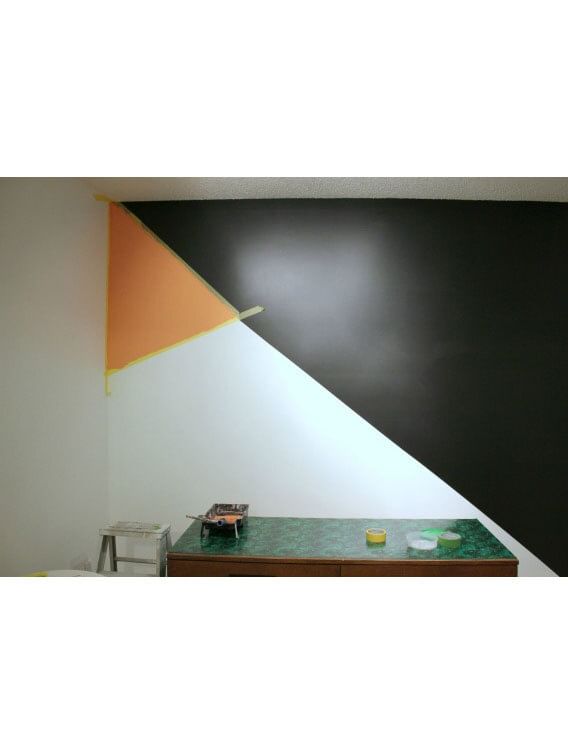 Geometric painted wall. Diagonal line splitting the wall in half. Upper left small orange triangle, bottom left white, right side black. Small green table at the bottom of the image