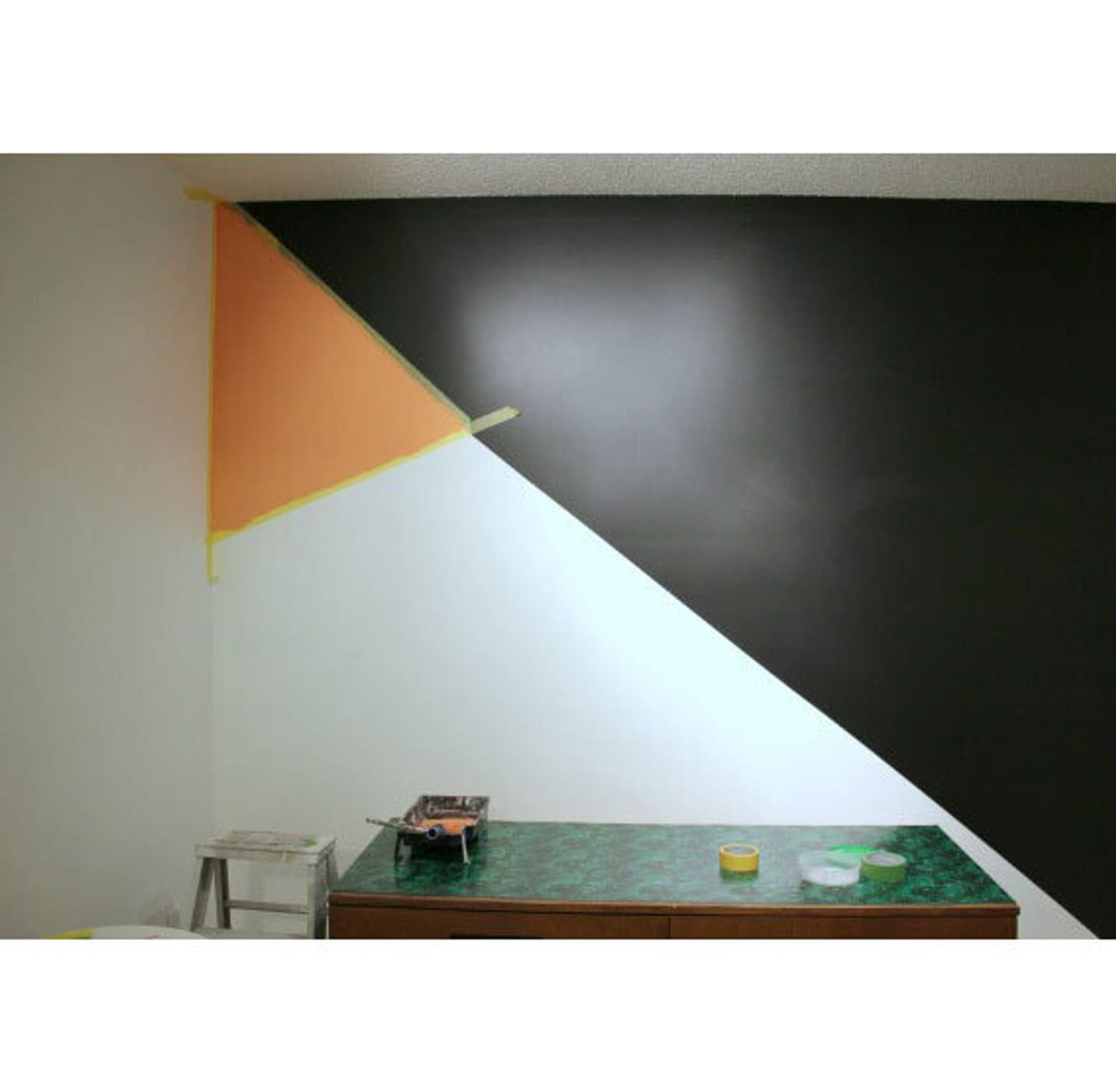 Geometric painted wall. Diagonal line splitting the wall in half. Upper left small orange triangle, bottom left white, right side black. Small green table at the bottom of the image