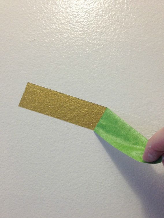 Green FrogTape Multi-Surface Painter's Tape being peeled off of a white wall to reveal a small crisp gold rectangle