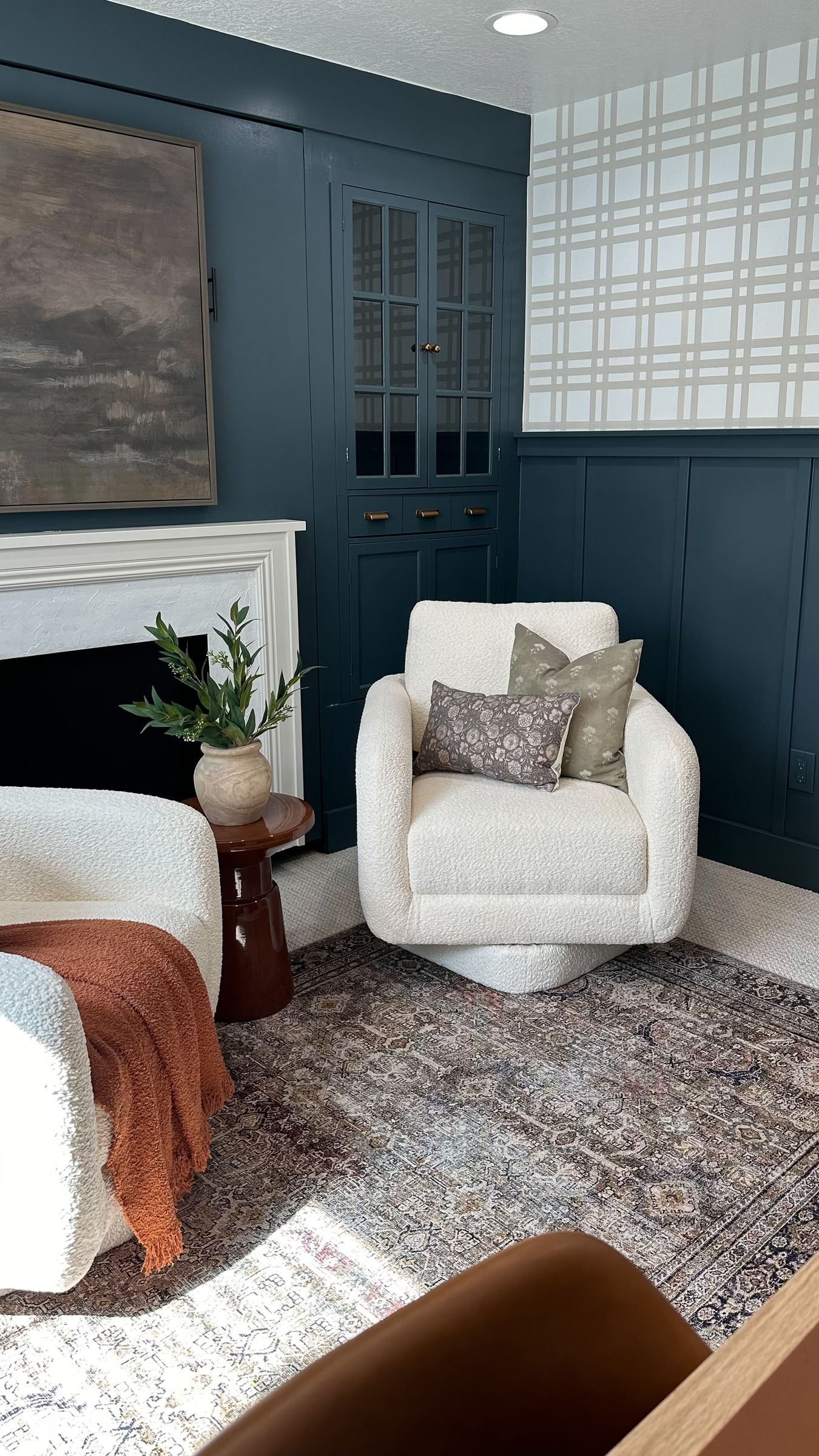 Cozy living room with cream swivel chairs, patterned cushions, a brown throw, and a potted plant. Blue walls, plaid wallpaper, and a large painting.