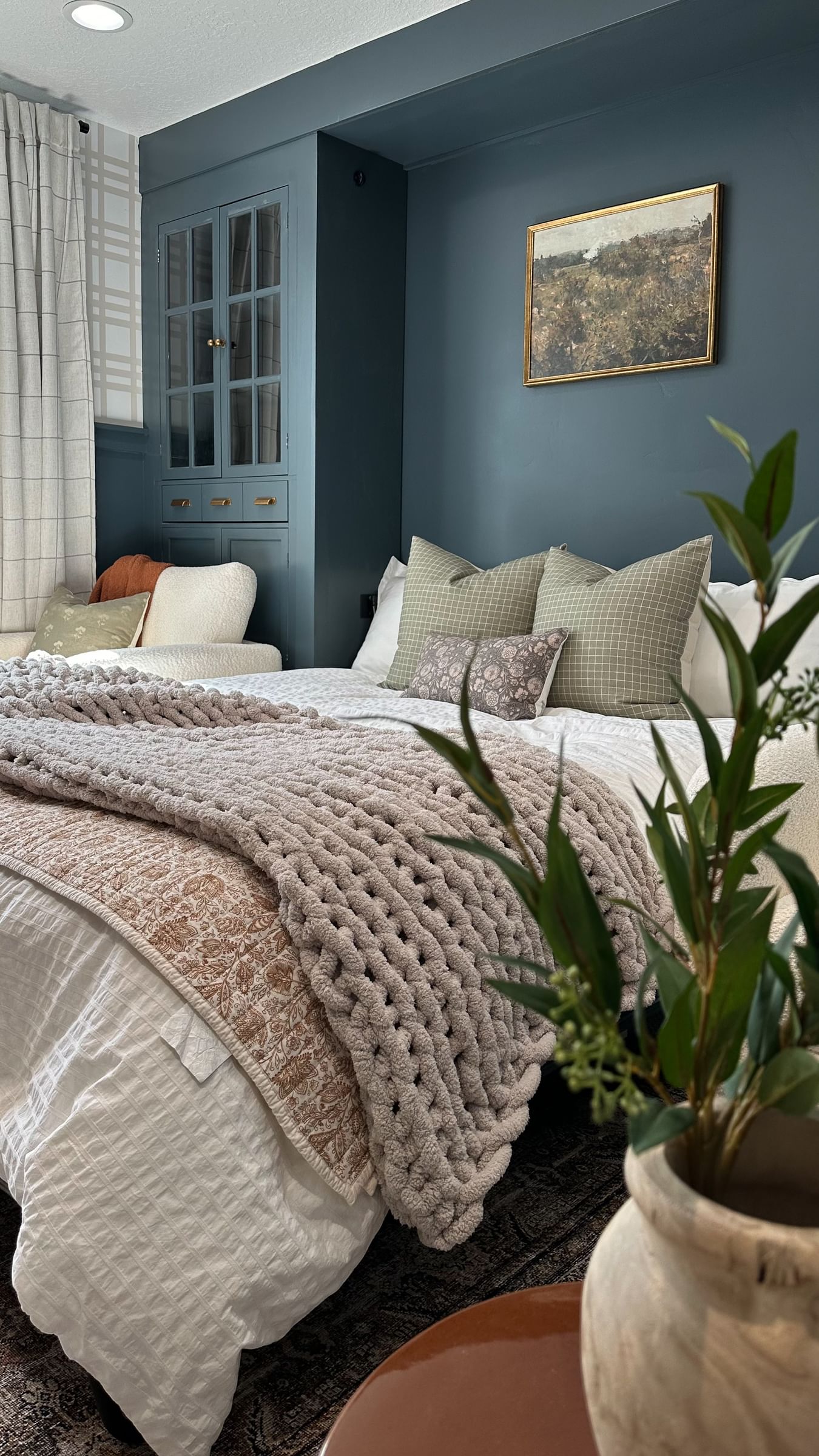 Cozy bedroom with teal walls, a white bed adorned with textured pillows and a thick knit gray blanket, a potted plant, and a landscape painting.