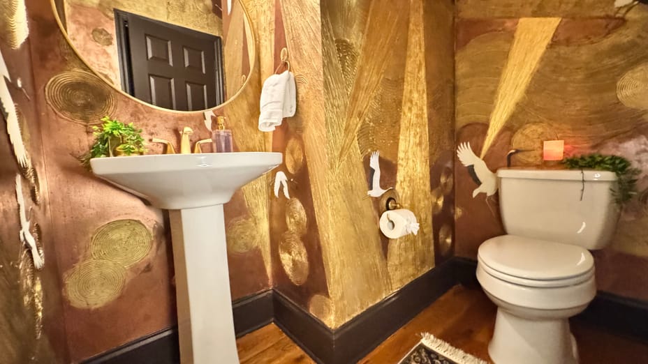 Luxurious bathroom with gold and brown walls, adorned with bird motifs. A round mirror hangs above a pedestal sink with gold fixtures and a plant. To the right, a toilet sits on warm wooden flooring, complemented by a patterned rug. The ambiance is elegant and artistic.