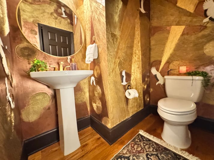 Luxurious bathroom with gold and brown walls, adorned with bird motifs. A round mirror hangs above a pedestal sink with gold fixtures and a plant. To the right, a toilet sits on warm wooden flooring, complemented by a patterned rug. The ambiance is elegant and artistic.
