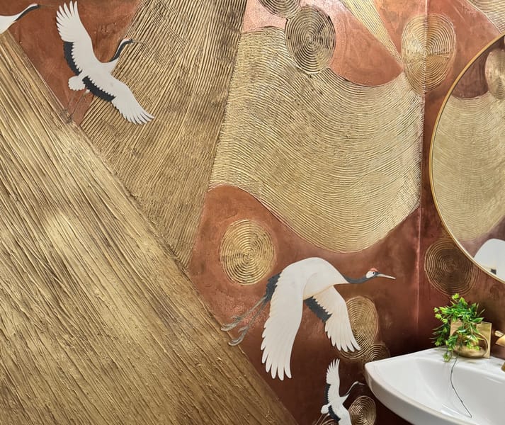 Elegant bathroom wall features gold and bronze textured patterns with white cranes in flight. A round mirror and a small plant above the sink add charm.