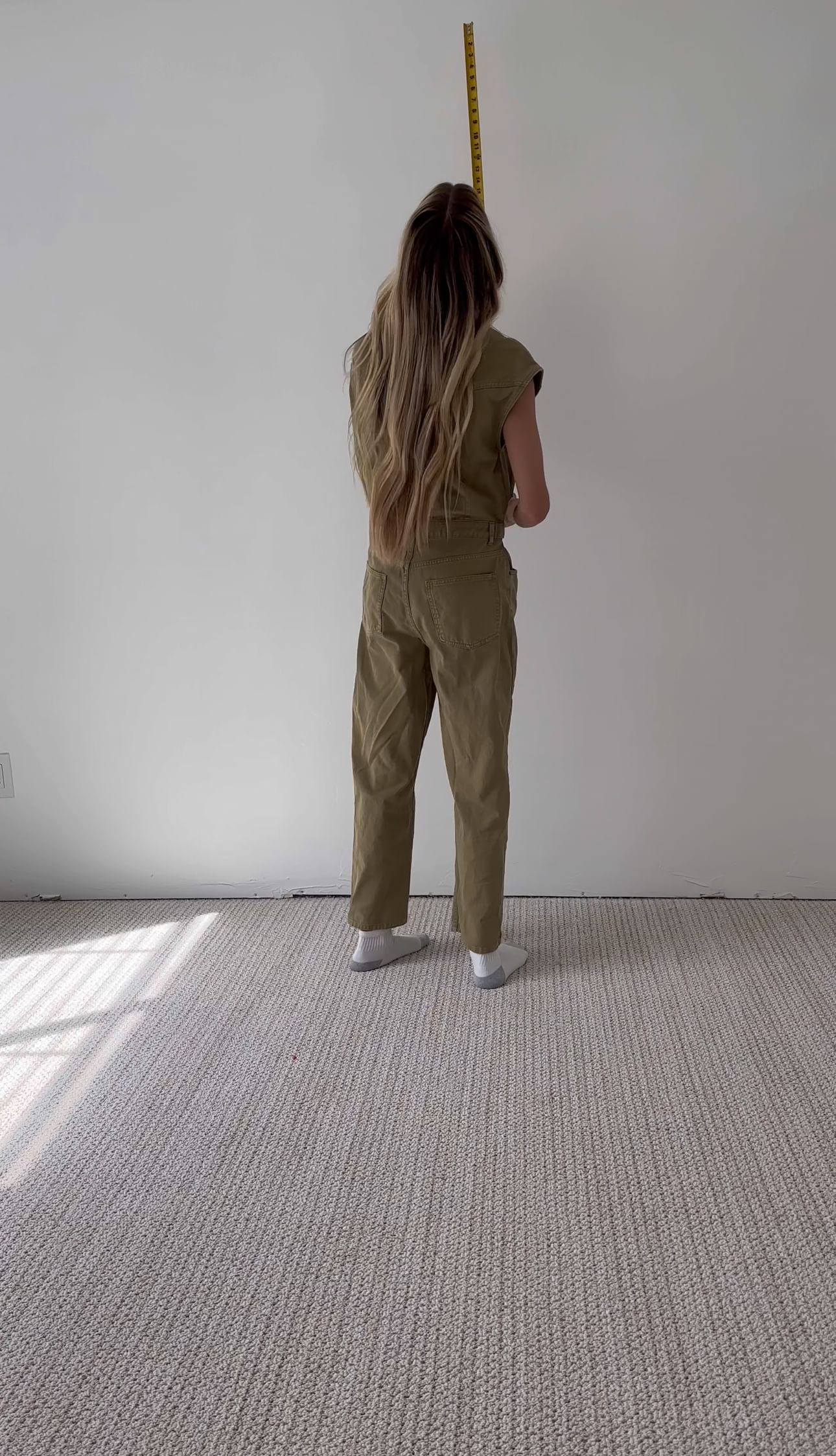 A person with long hair wearing a brown jumpsuit stands on a beige carpet, facing a white wall, holding a yellow tape measure vertically.