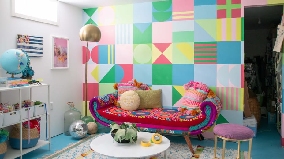 Room with a colorful wall mural. The mural consists of many multicolored green, blue, pink, and yellow squares with different shapes in them. There is a small colorful couch, stool, coffee table, lamp, and shelves.