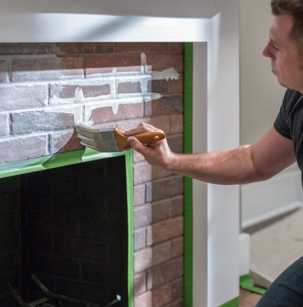 Man Whitewashing a brick fireplace using a paintbrush. The edges of the brick are masked off with Green FrogTape Multi-Surface Painter's Tape