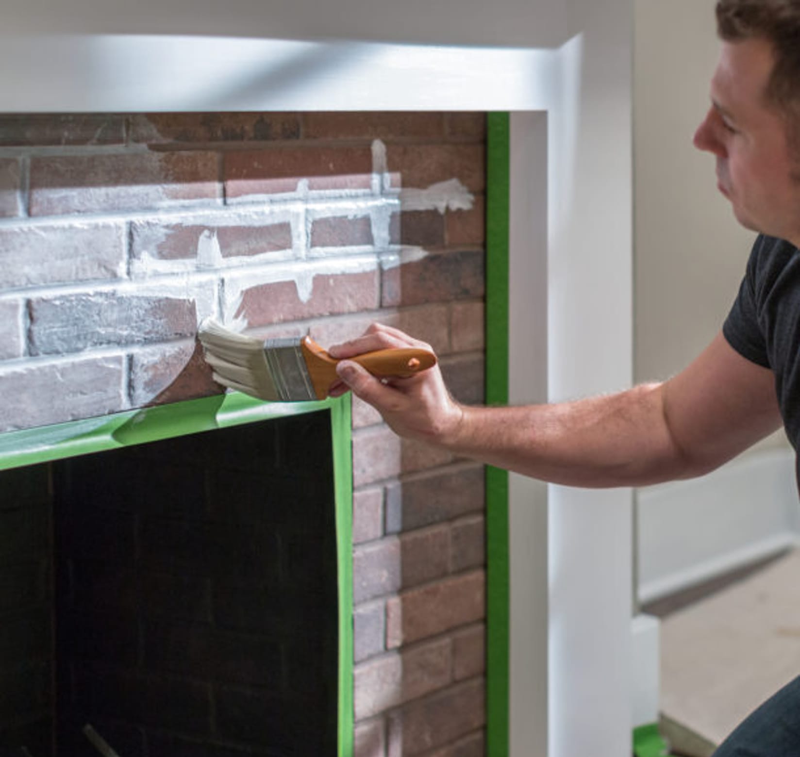 Man Whitewashing a brick fireplace using a paintbrush. The edges of the brick are masked off with Green FrogTape Multi-Surface Painter's Tape