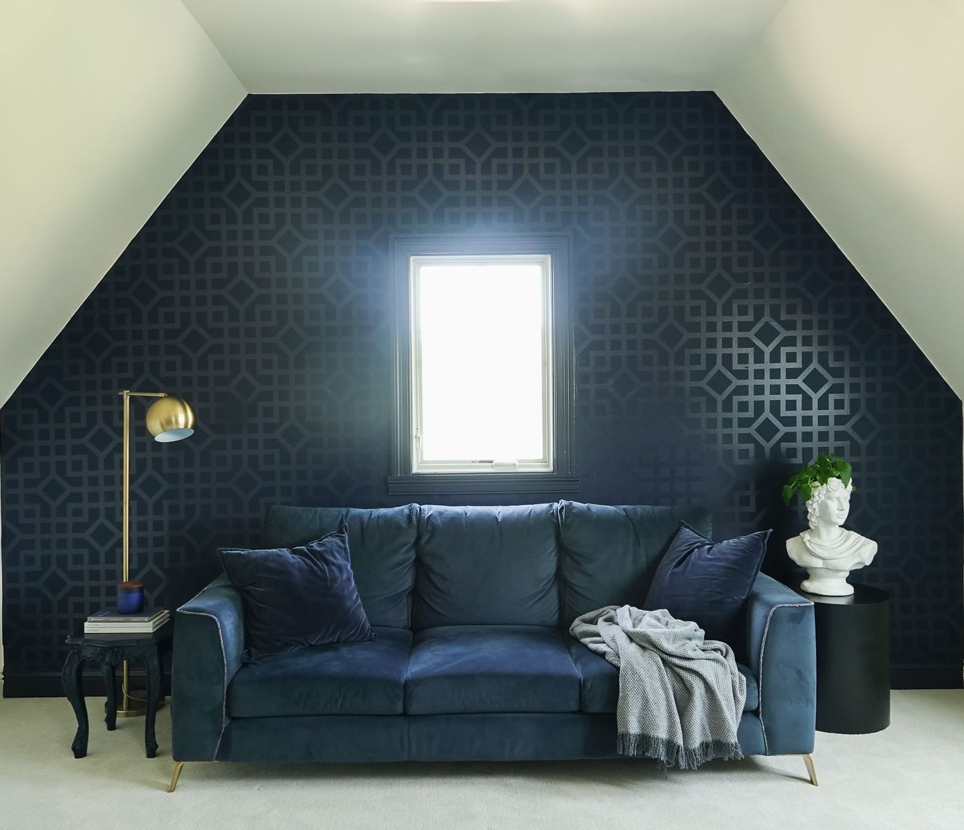 Dark Blue wall with intricate geometric pattern and rectangular window. Ther is a dark blue couch under the window with a gold lamp on the left and a white stone bust on the right.