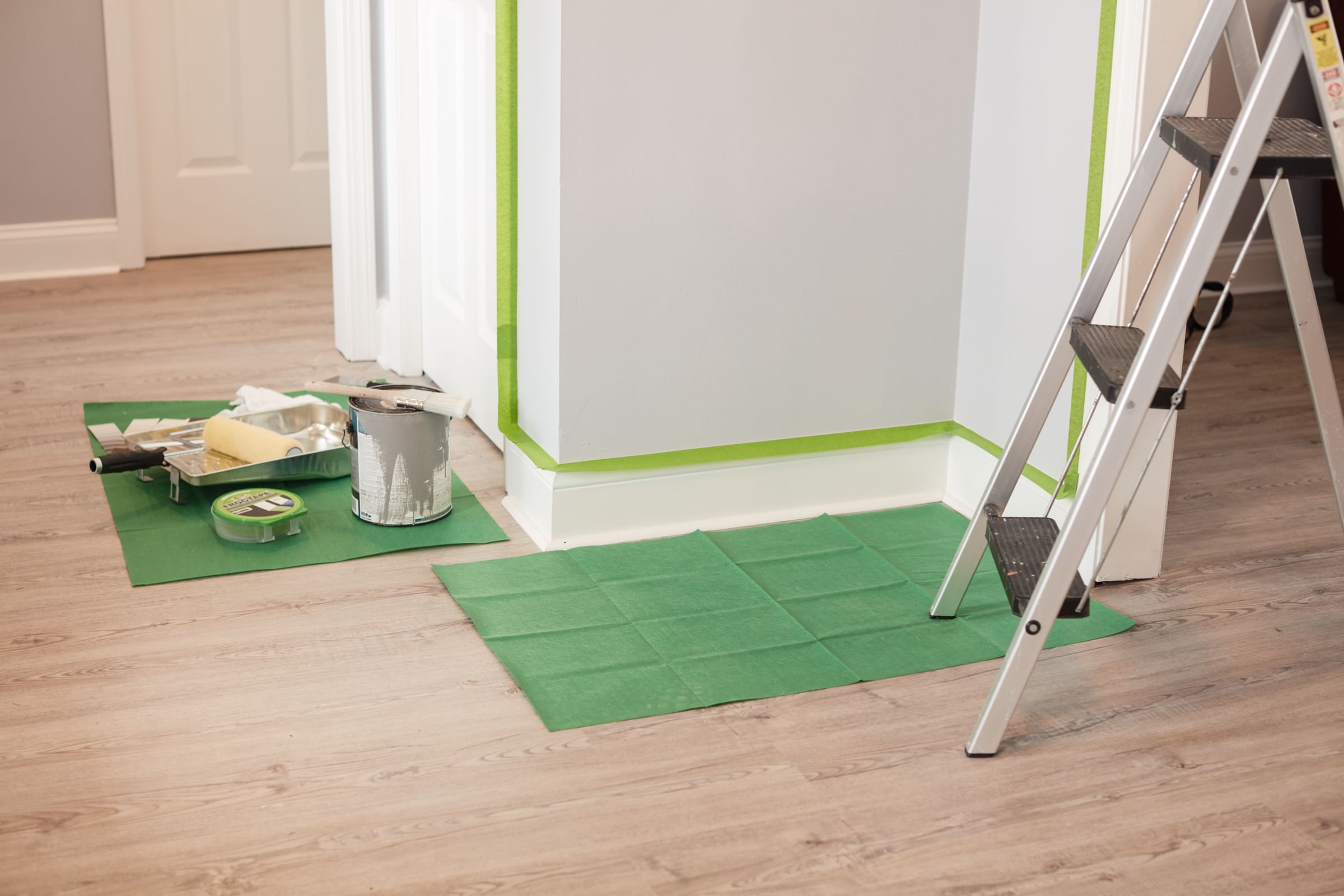 A paint site in a home, with Green FrogTape Multi-Surface Painter's Tape masking off the white trim. There are green drop cloths on the floor, a paint can, and a step ladder.