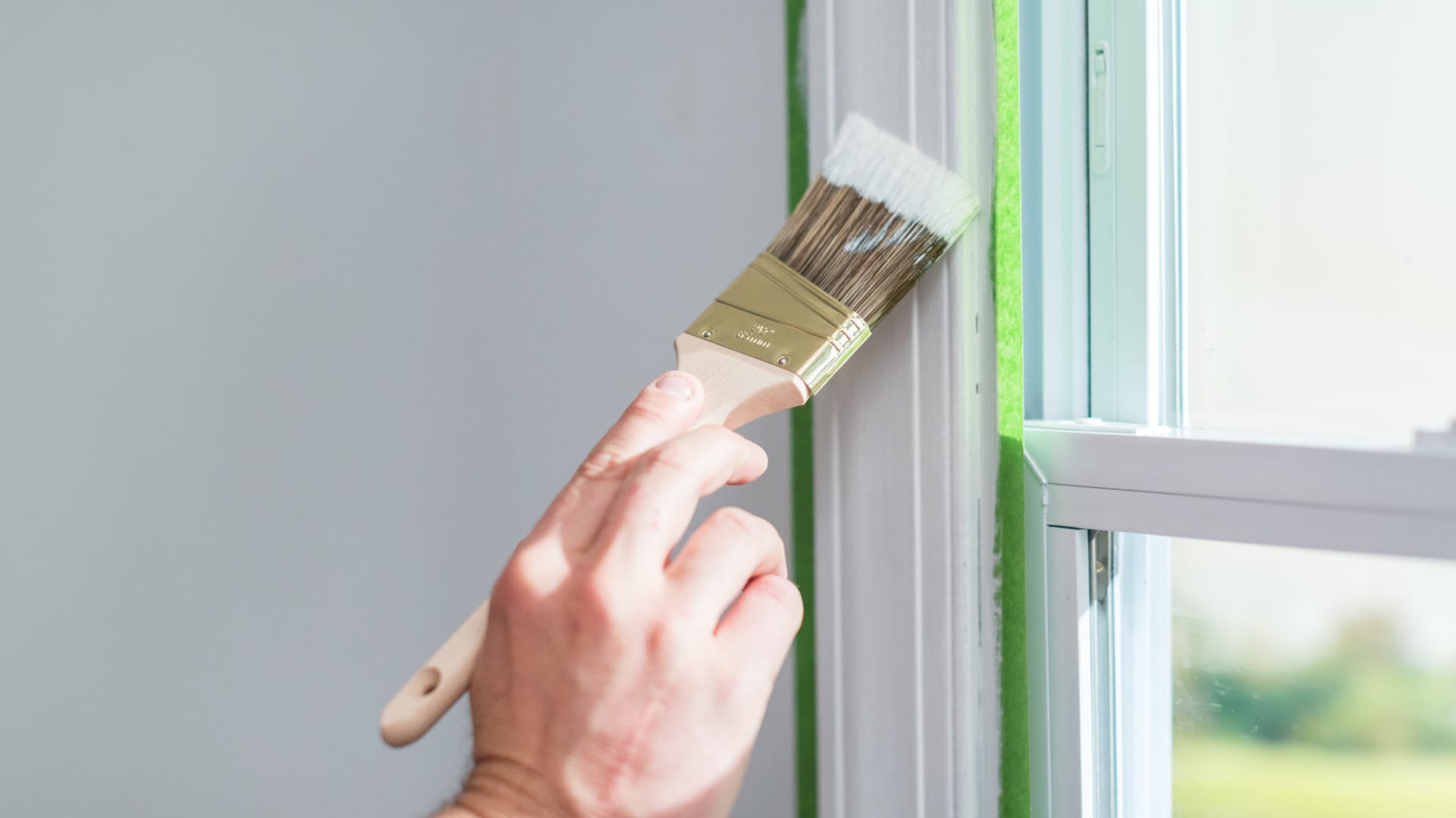 A hand using a paintbrush to paint window trim white. The window trim is taped off with Green FrogTape Multi-Surface Painter's Tape