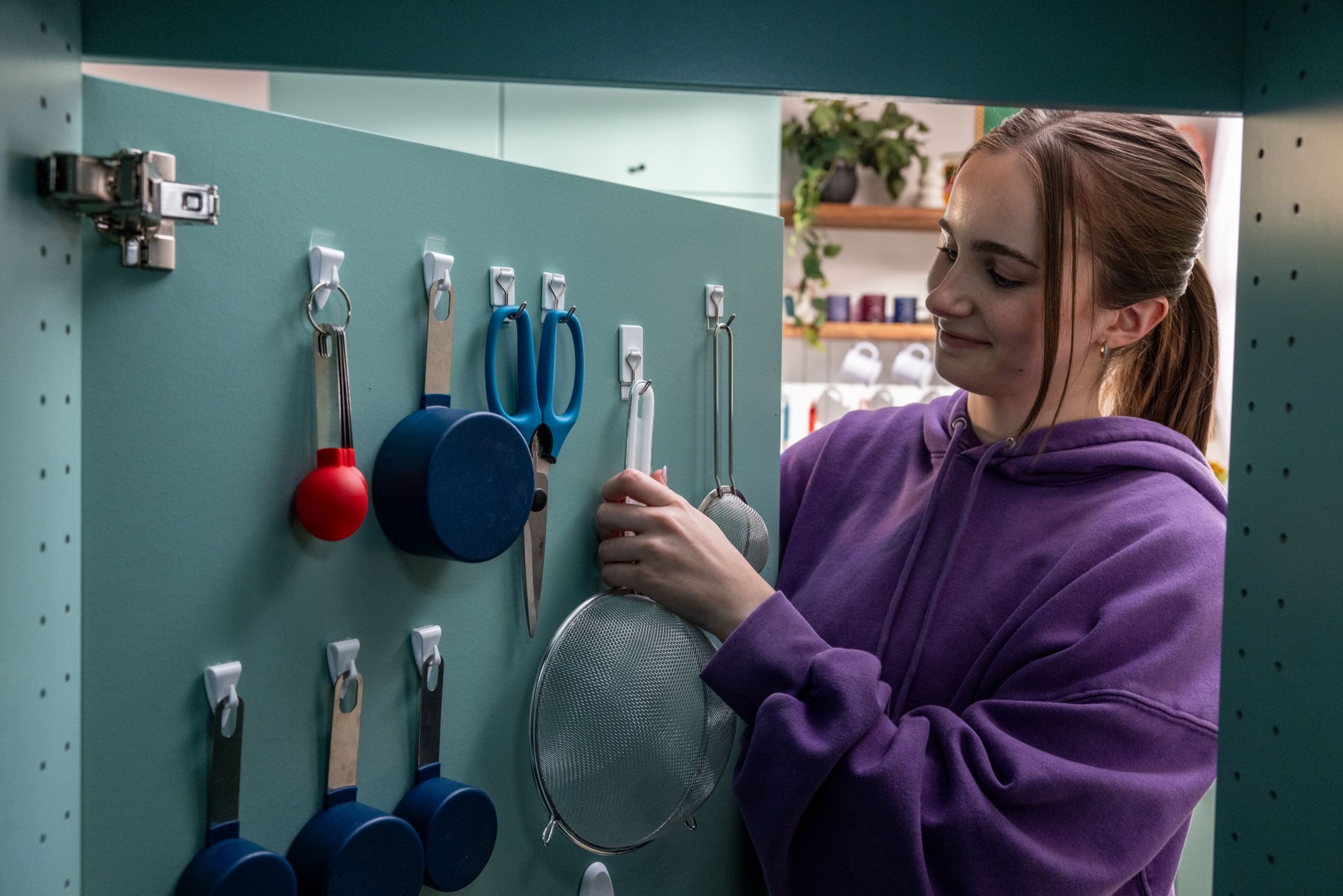 A woman hanging a strainer from a FrogTape reMOVEables hook on the back of a kitchen cabinet. There are other miscellaneous utensils also hanging on FrogTape reMOVEables hooks on the back of the cabinet.