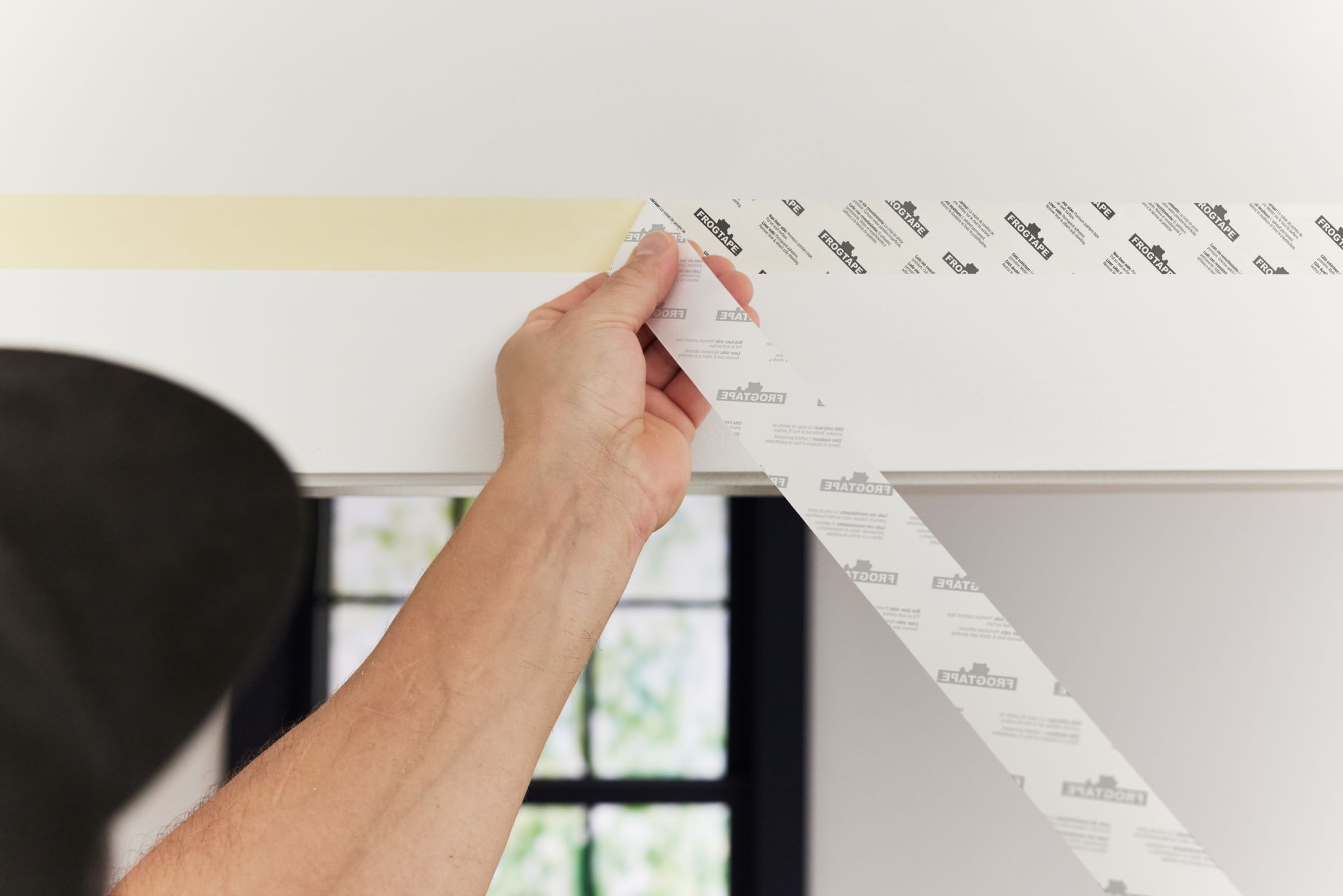 A man peeling the adhesive backing off of FrogTape DS154. The Tape is applied to the top of a doorway.