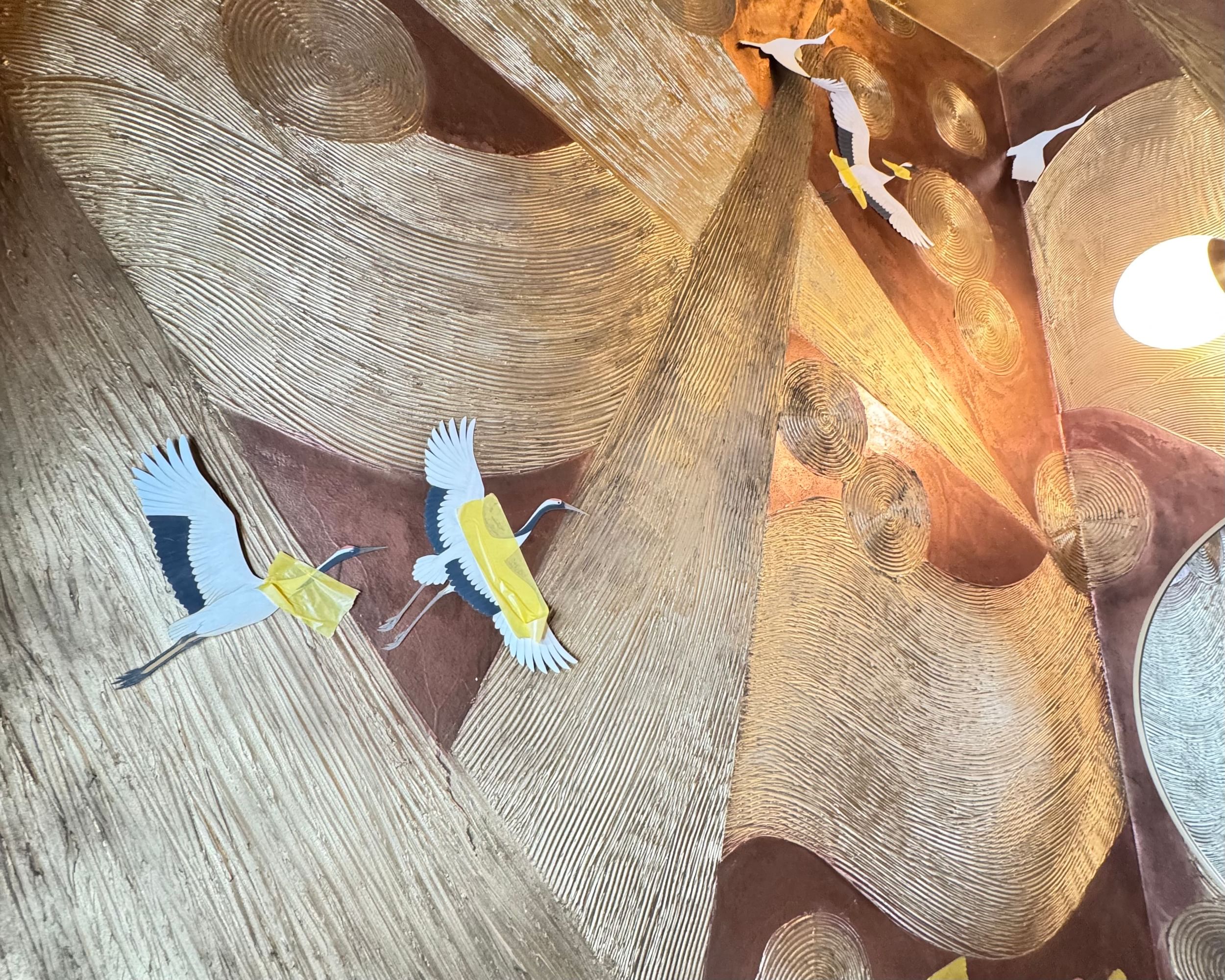 Textured brown wall art with swirling, circular patterns and vibrant, paper-crafted cranes in flight. A warm, ambient light adds a serene atmosphere.