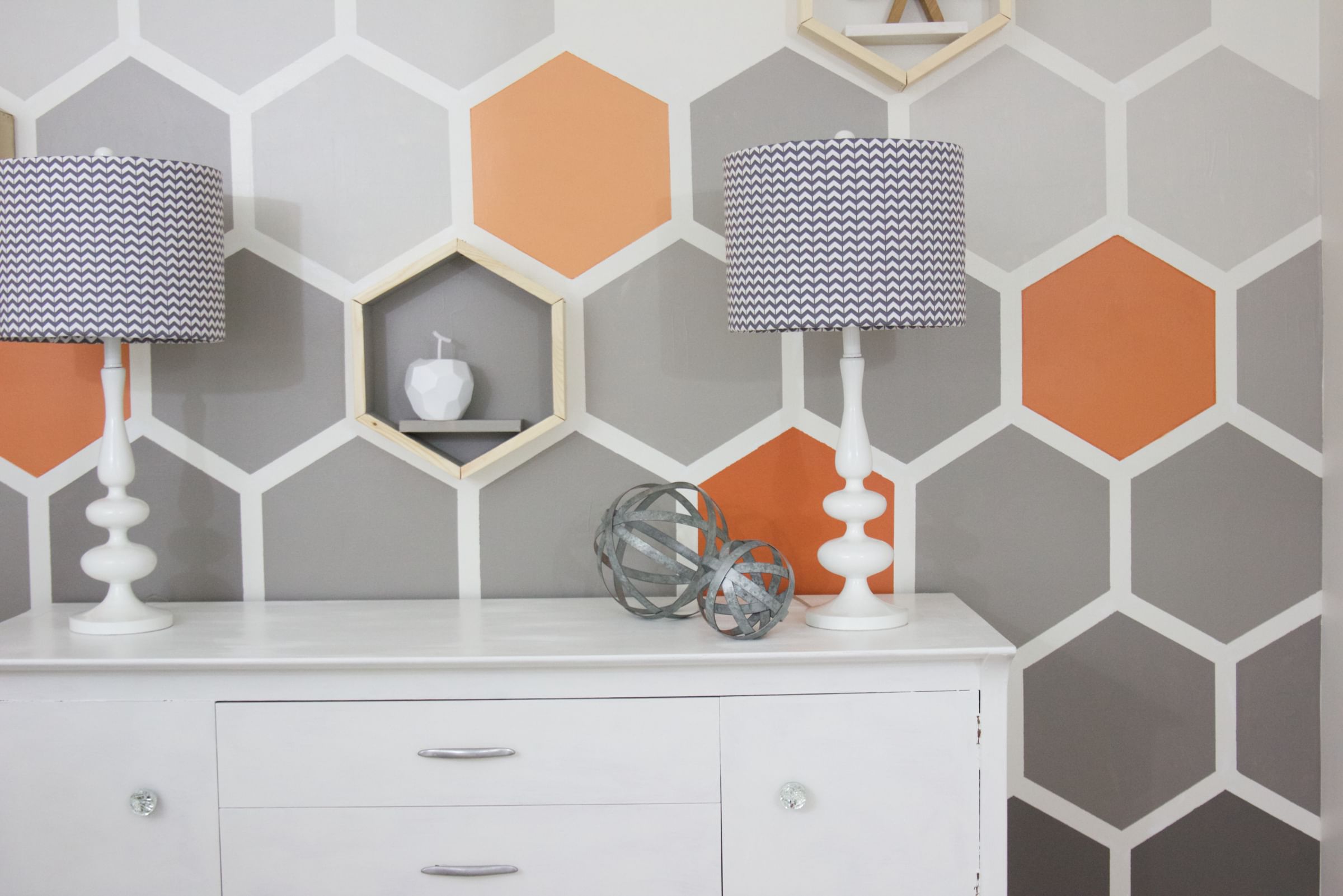 Orange and gray honeycomb pattern on a wall with a small white dresser with two lamps on it in front
