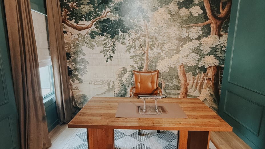 Elegant office with a wooden desk and leather chair. The room features a forest-themed mural, green walls, and a checkered rug, creating a serene ambiance.