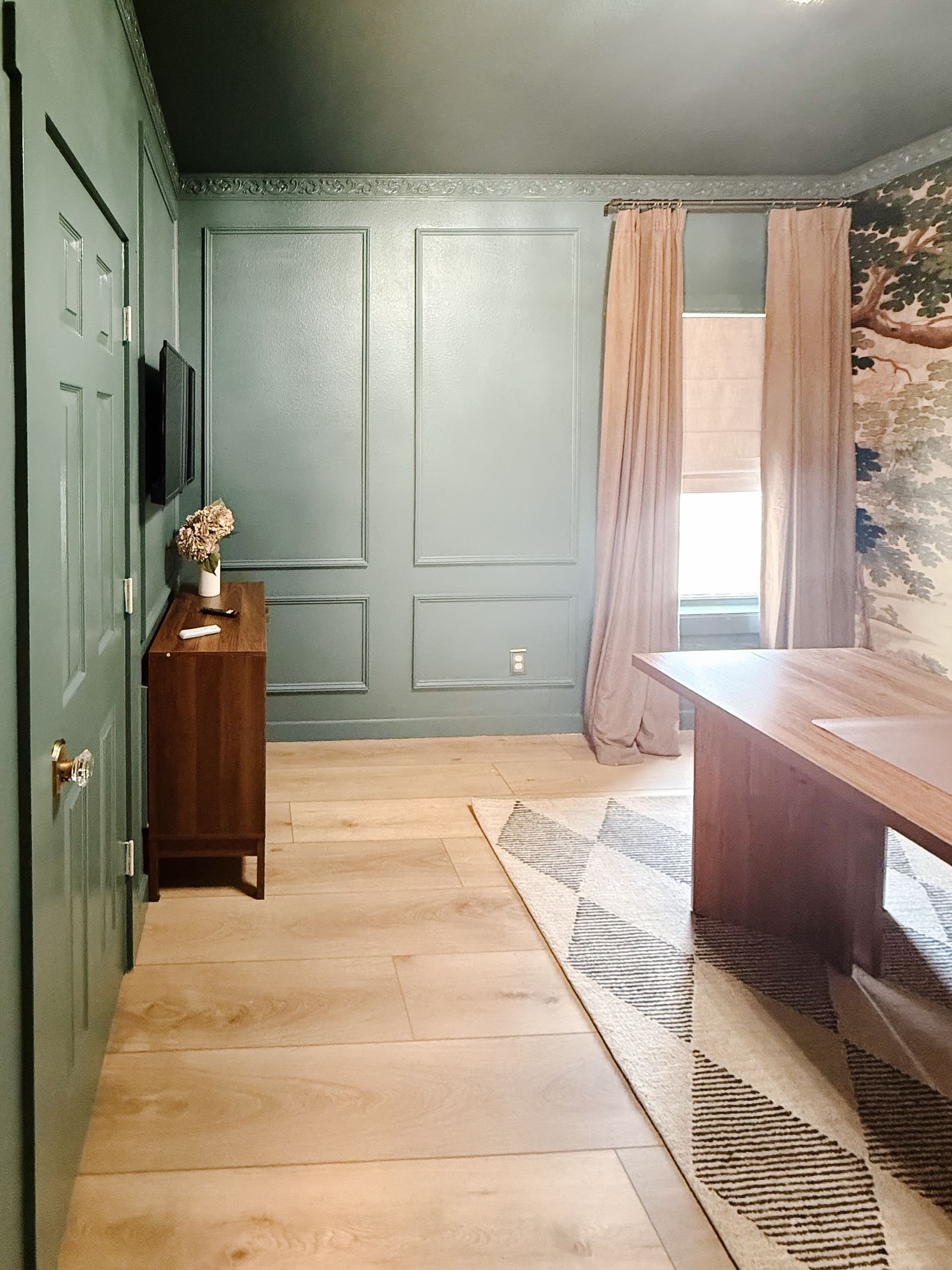 Stylish room with green walls and molding, wood floor, and soft lighting. Features a wooden desk, pink curtains, floral wallpaper, and a chevron rug.