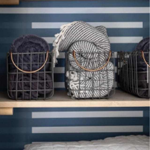 Three metal baskets on a wooden shelf with blankets in them. The back wall is back blue with alternating thin rectangle strips
