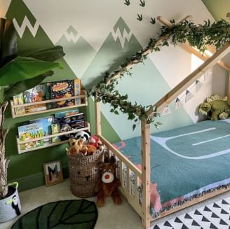 A child's bedroom with a green mountain mural on the wall. There are two small wall book shelves and a toddler bed.