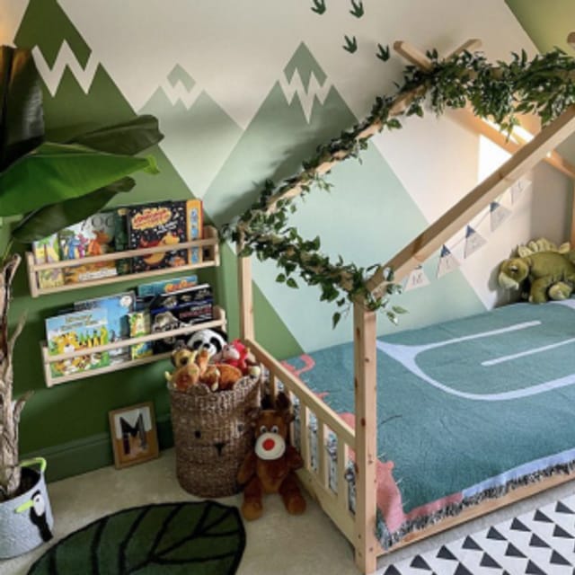 A child's bedroom with a green mountain mural on the wall. There are two small wall book shelves and a toddler bed.
