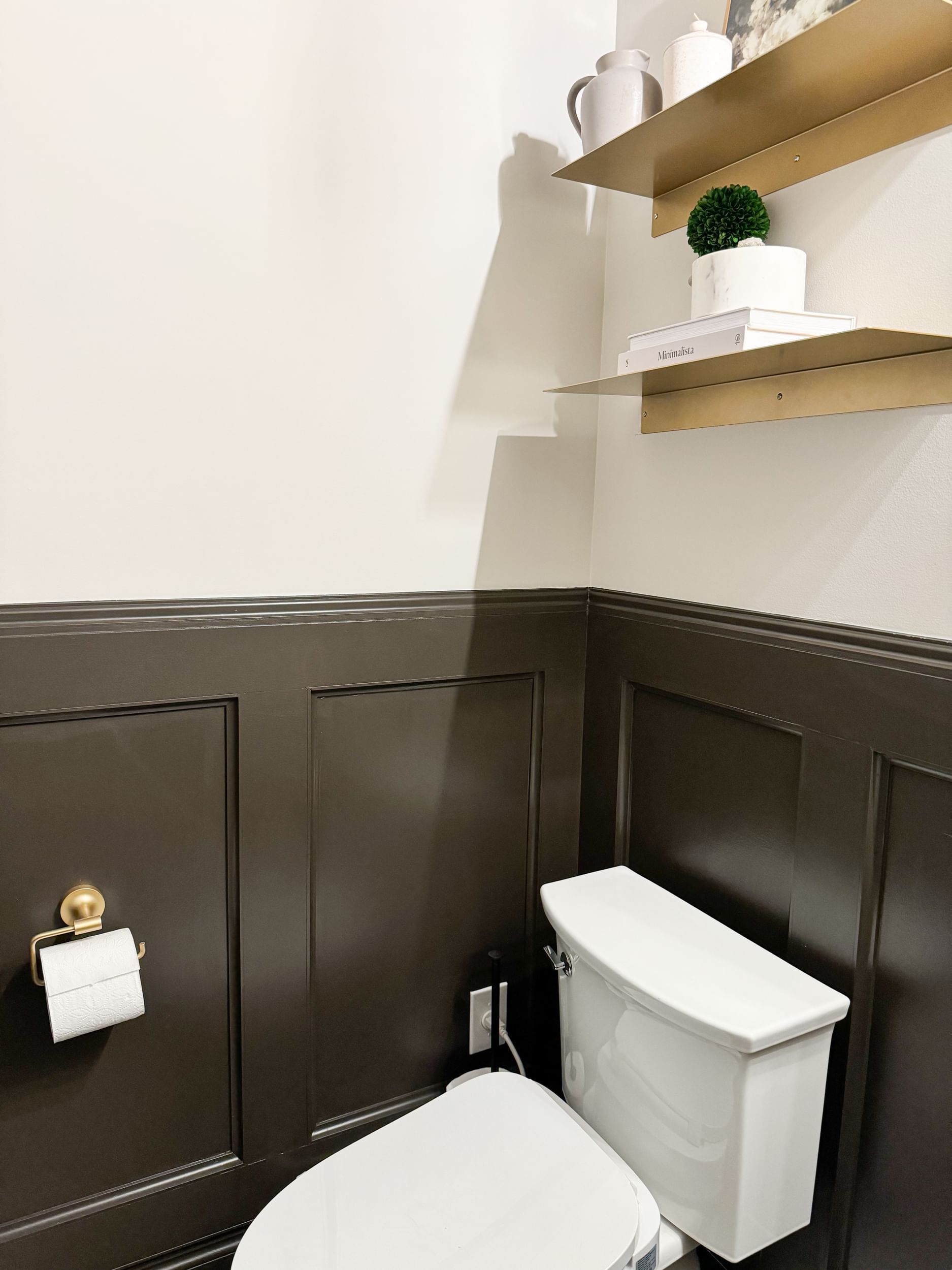 Bathroom with Brown Wainscotting. There are two gold shelves on a cream colored wall above the toilet.