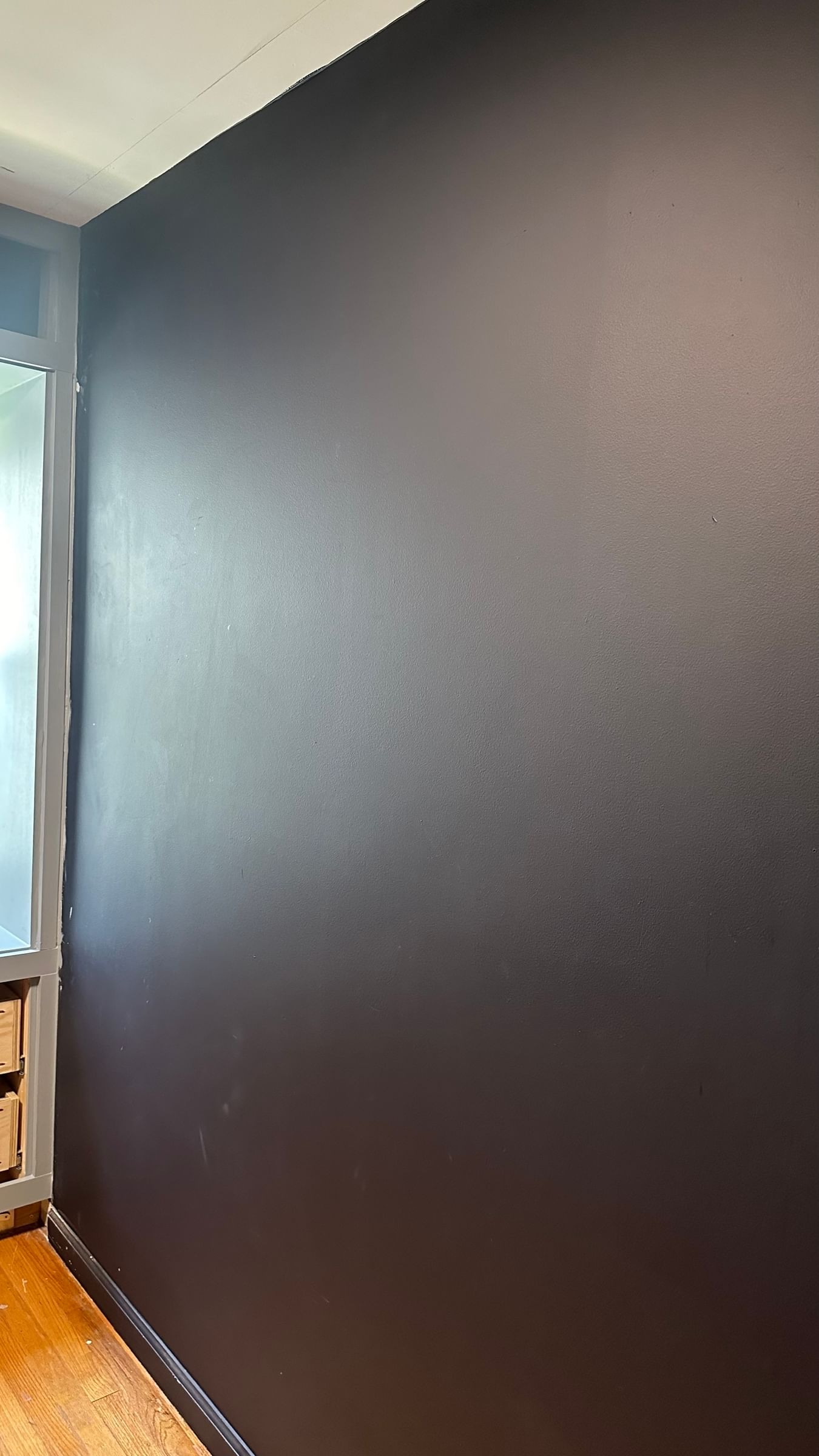 Smooth black accent wall in a room, contrasting with adjacent white ceiling and wooden floor. A window partially visible on the left adds light.