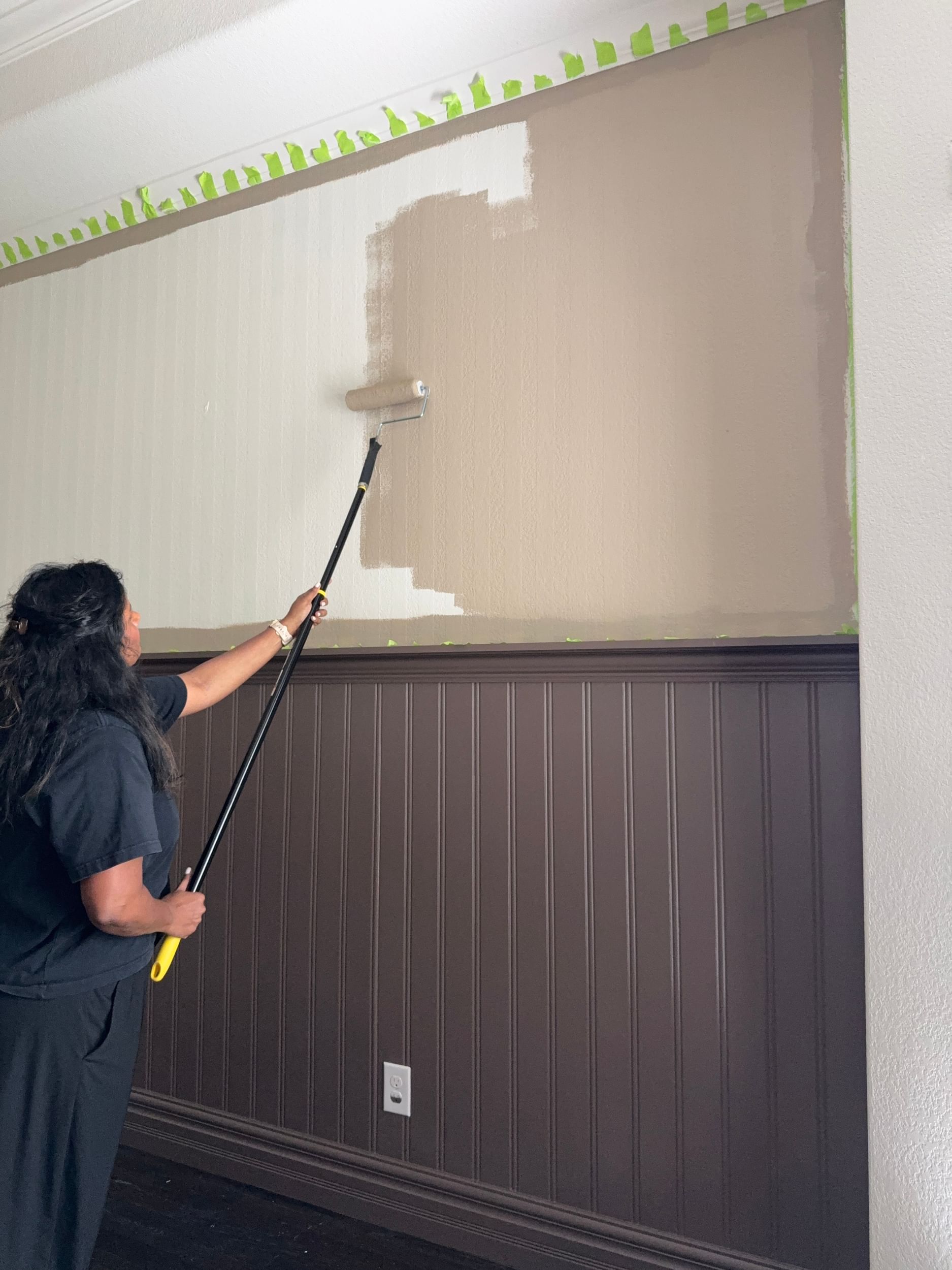Light brown paint being applied to the top section of a wall with a roller. The wall has vertical stips of FrogTape Advanced