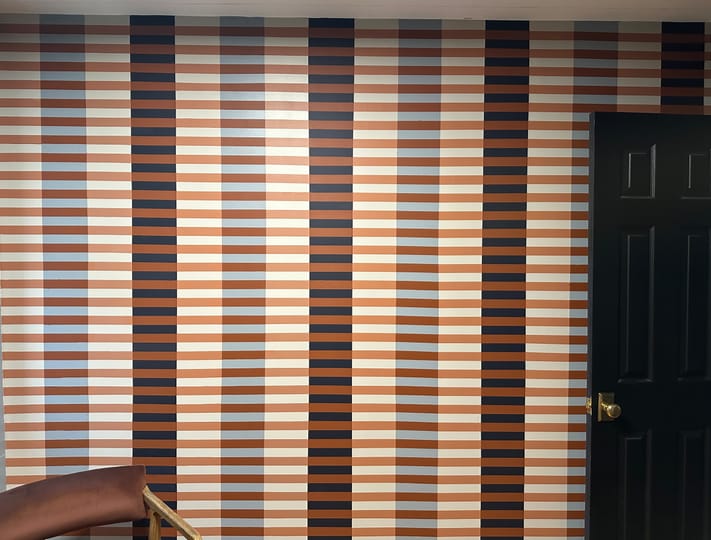 Striped wall art with vertical bands in brown, orange, cream, and blue creates a retro feel. A black door is on the right, and part of a brown chair is visible on the left.